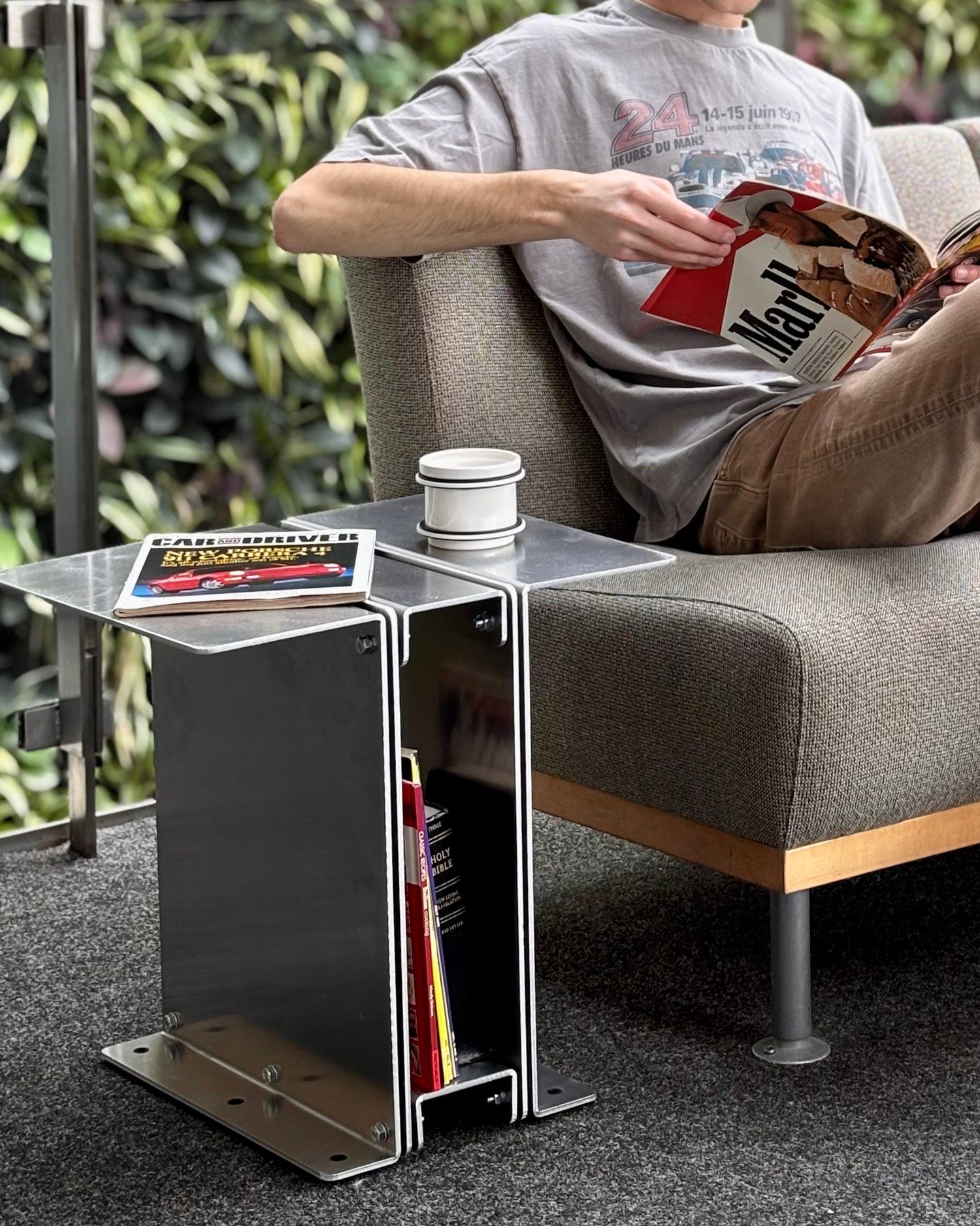 Someone reading on a sofa with the 3082 Book Table beside them, holding books and a mug